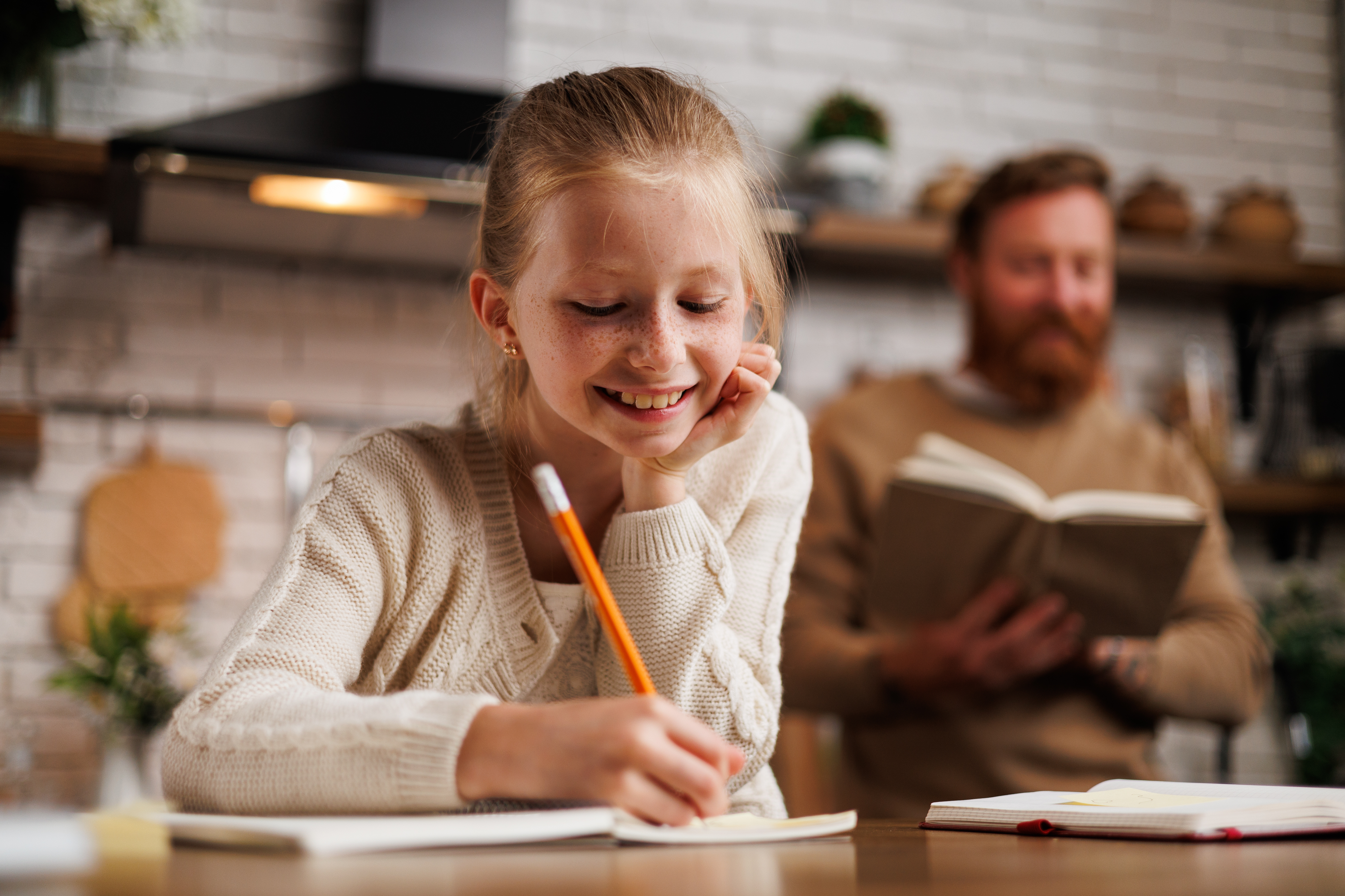 Female student sitting at kitchen counter working on assignment while adult male stands reading a book behind her