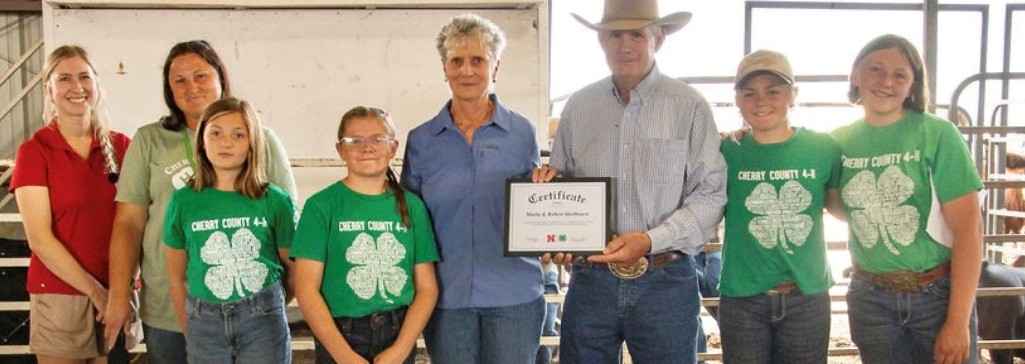 A group of people, some wearing 4-H clover shirts, stand indoors holding a certificate.
