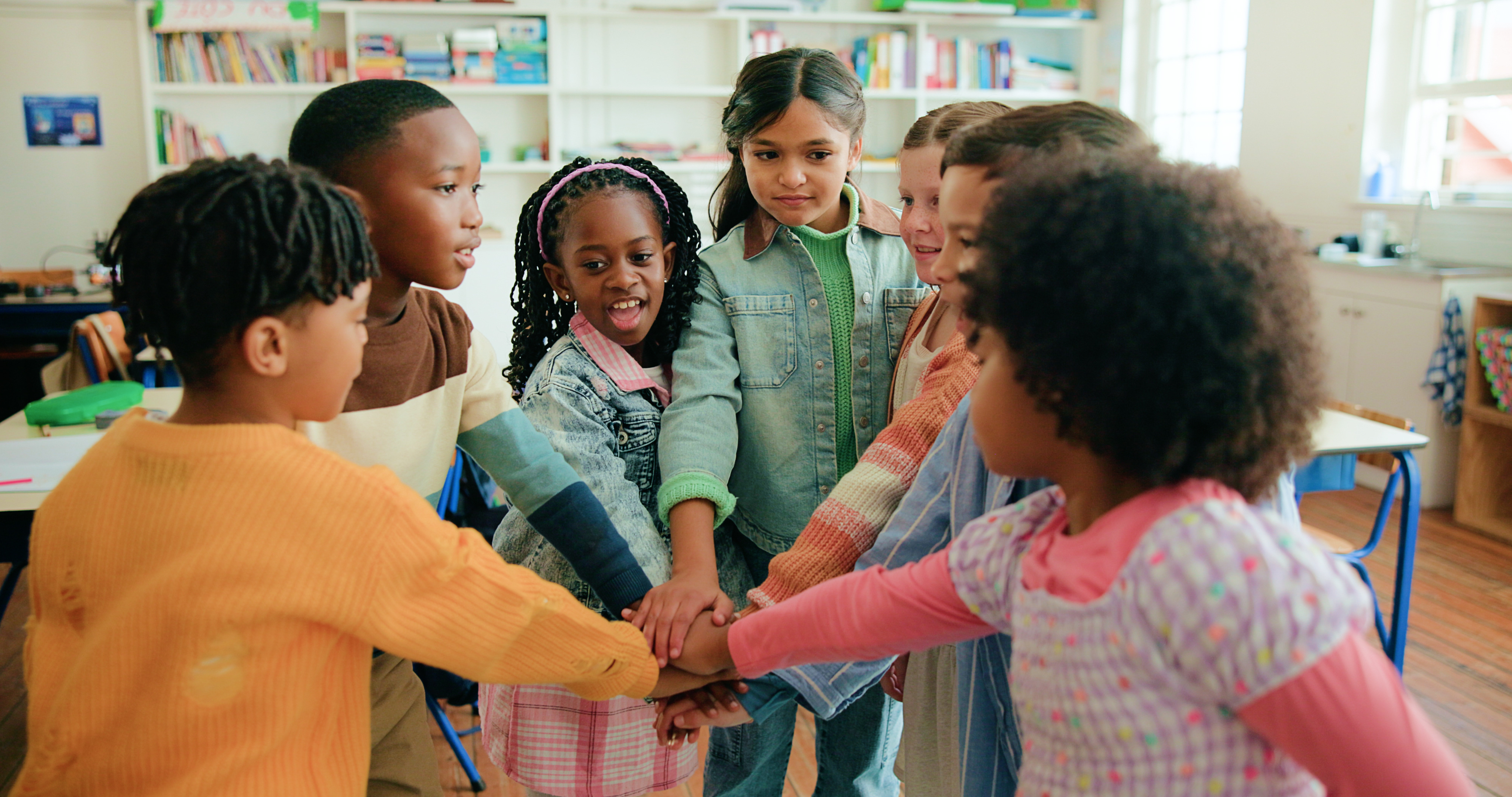 Seven students standing in a circle with their hands in the middle inside of a classroom