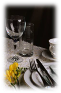 Elegant table setting with glassware, a white teacup, cutlery, and yellow flowers on a lace tablecloth.