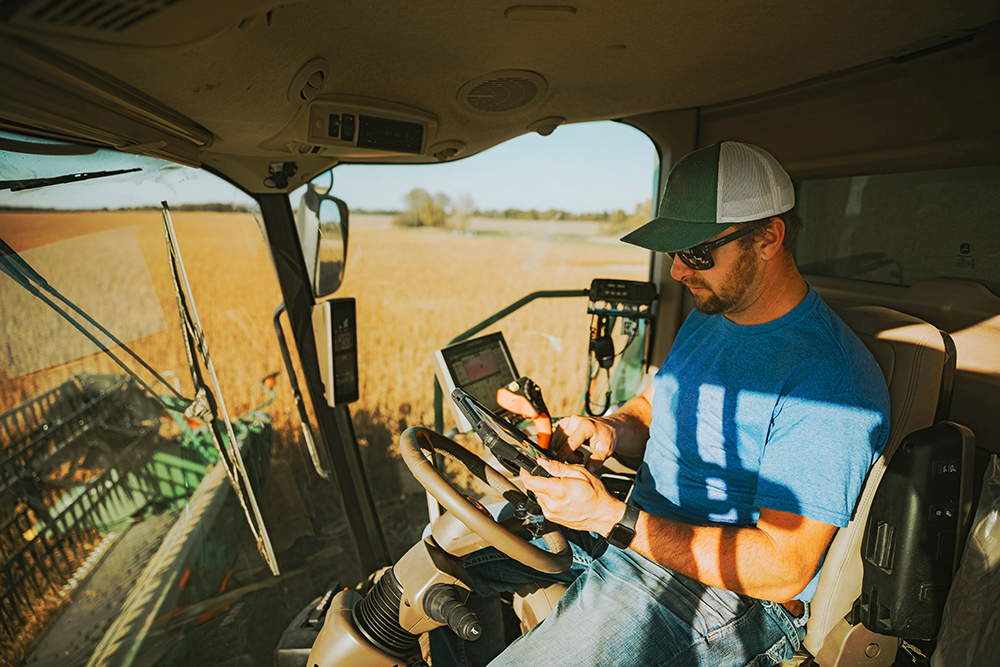 Farmer in a tractor cab, using a smartphone while operating controls in a sunlit field.