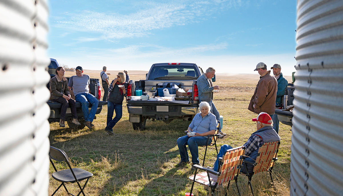A group of people socializing near pickup trucks in a sunny field, two silos partially framing the scene.