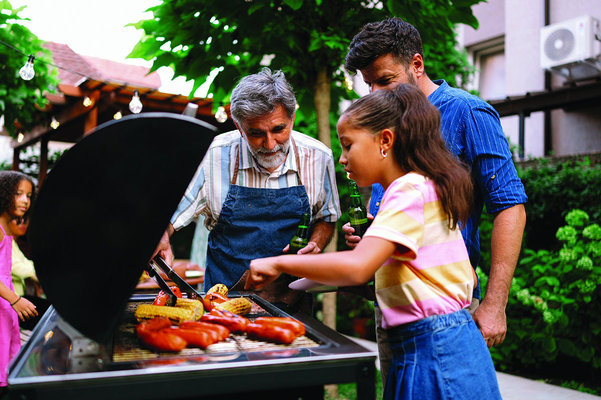 Family grilling sausages and corn together outdoors in a backyard setting.