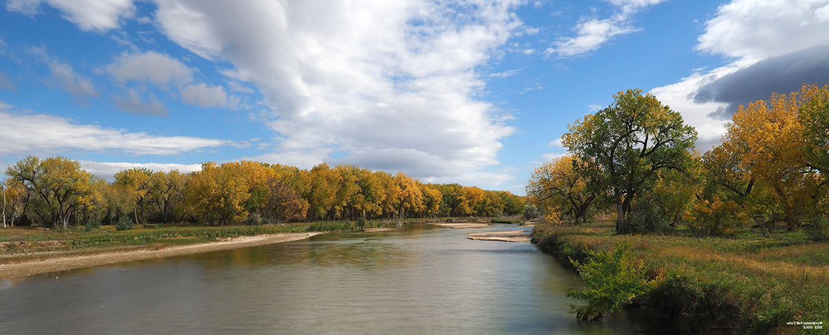 A calm river with trees in autumn colors under a partly cloudy blue sky.
