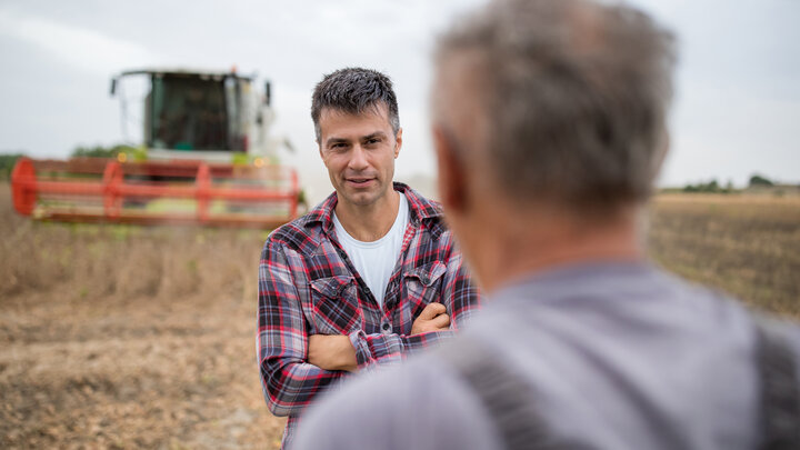 Two male farmers — one younger, one older — talk in a soybean field with a combine in the background.