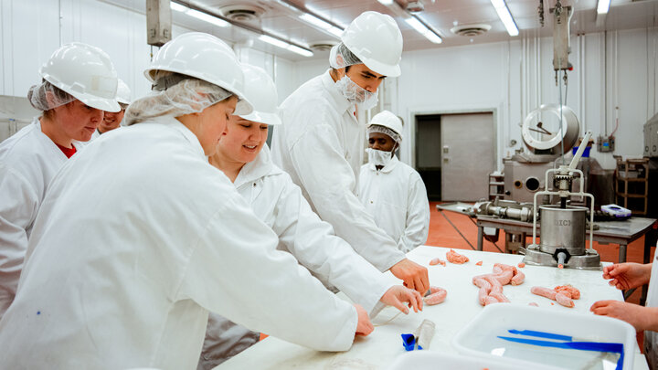 Students in the UNL Meat Science Club — wearing labcoats, hair nets and white hard hats — work with a sausage maker. Several sausages lie on the table.