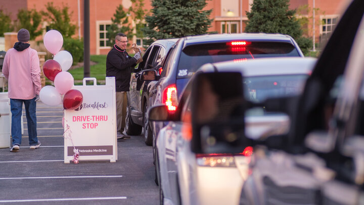 A man hands a breakfast bag through a car window in a parking lot. Several other vehicles are lined up behind the car.