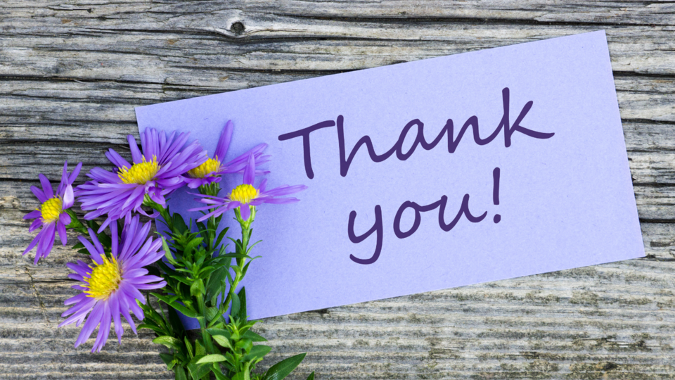 A purple note that says thank you with a bouquet of purple asters next to it on a wooden surface.
