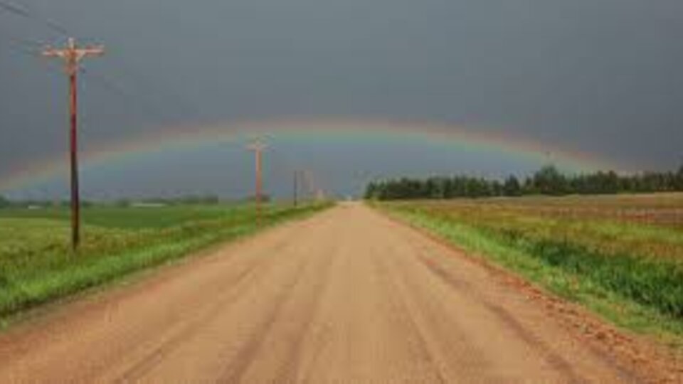 Dirt road under a stormy sky with a rainbow arching above fields and power lines.