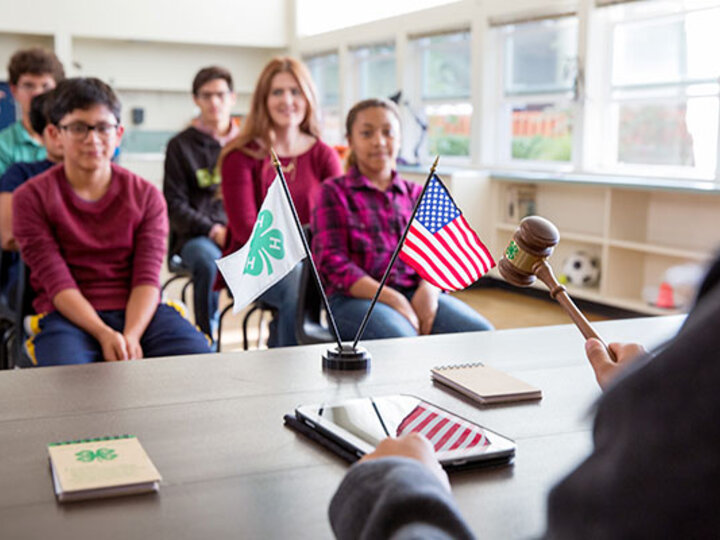 Youth 4-H Club members sitting in rows behind a table with a 4-H and U.S. flag with a club President holding a gavel