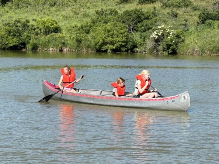 Three youth in a canoe on the lake with green brush in the background
