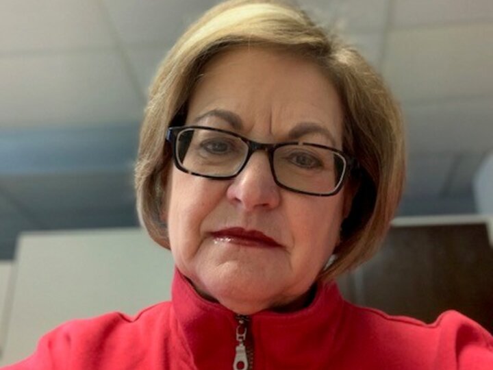 Woman wearing glasses and a red zip-up shirt, seated indoors with a ceiling in the background.
