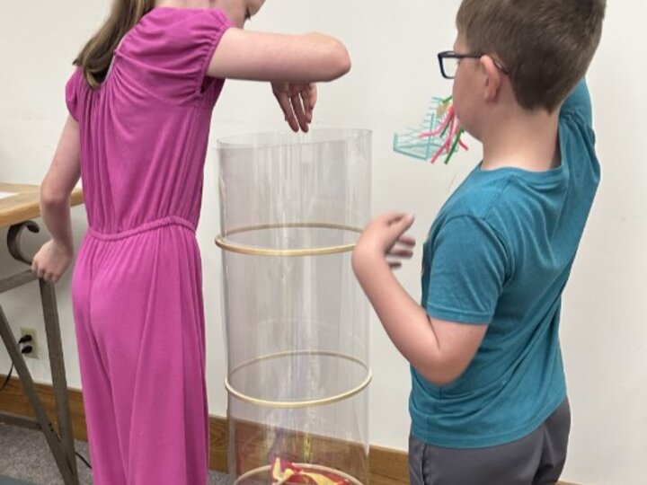 A girl wearing a pink jumpsuit drops her engineered contraption into a wind tunnel while a boy wearing a blue shirt holds his engineered contraption in the air and onlooks.