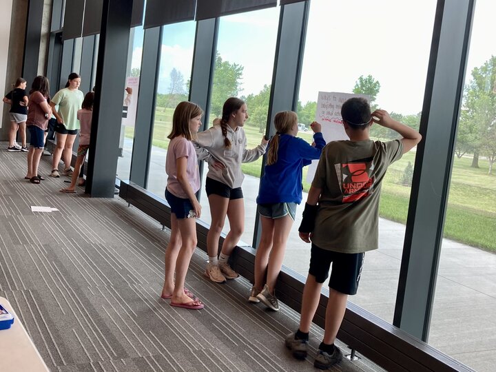 Students in casual attire stand by large windows reading posters, sunlight highlights a green lawn.