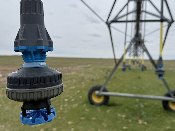 Irrigation nozzle in focus near a large sprinkler system on grassy farmland.