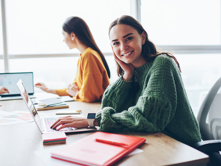 Smiling woman in a green sweater working on a laptop at a shared office table.