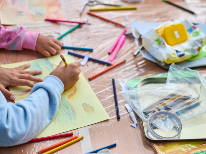 Children drawing with pencils on colorful paper amid scattered art supplies.