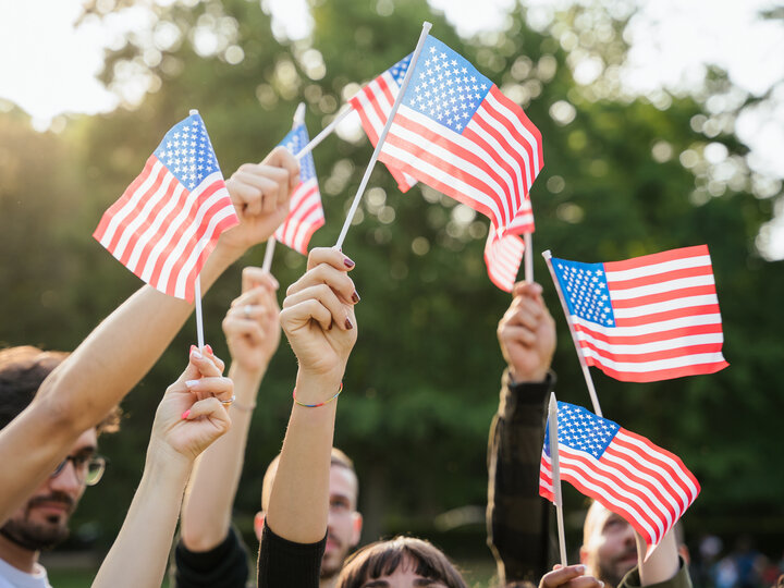 Hands waving small American flags outdoors with trees in the background.