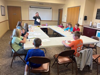 Library setting, multiple youth around a rectangular table with instructor at the top center instructing youth to cut playdough