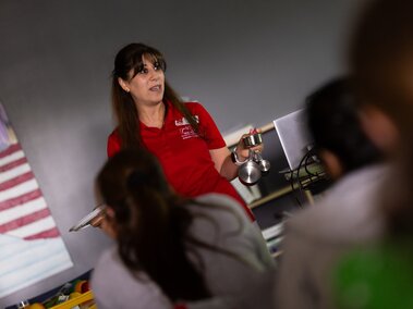 Instructor holds measuring cups, teaching a group in a classroom setting.