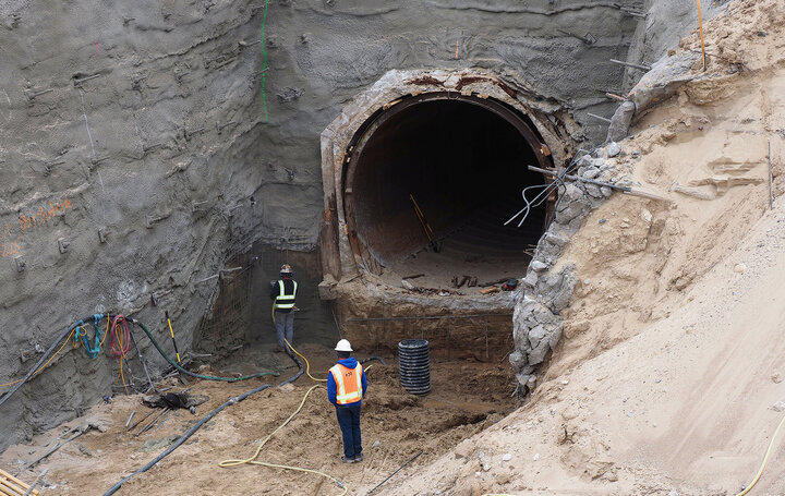 Construction workers near a large, partially-built tunnel entrance in a sandy excavation site.